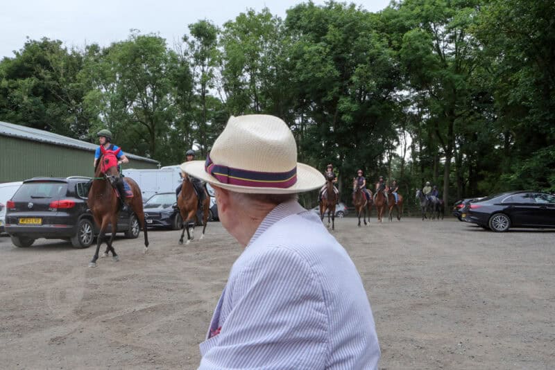 a man in a panama hat watches a string of racehorses ride past
